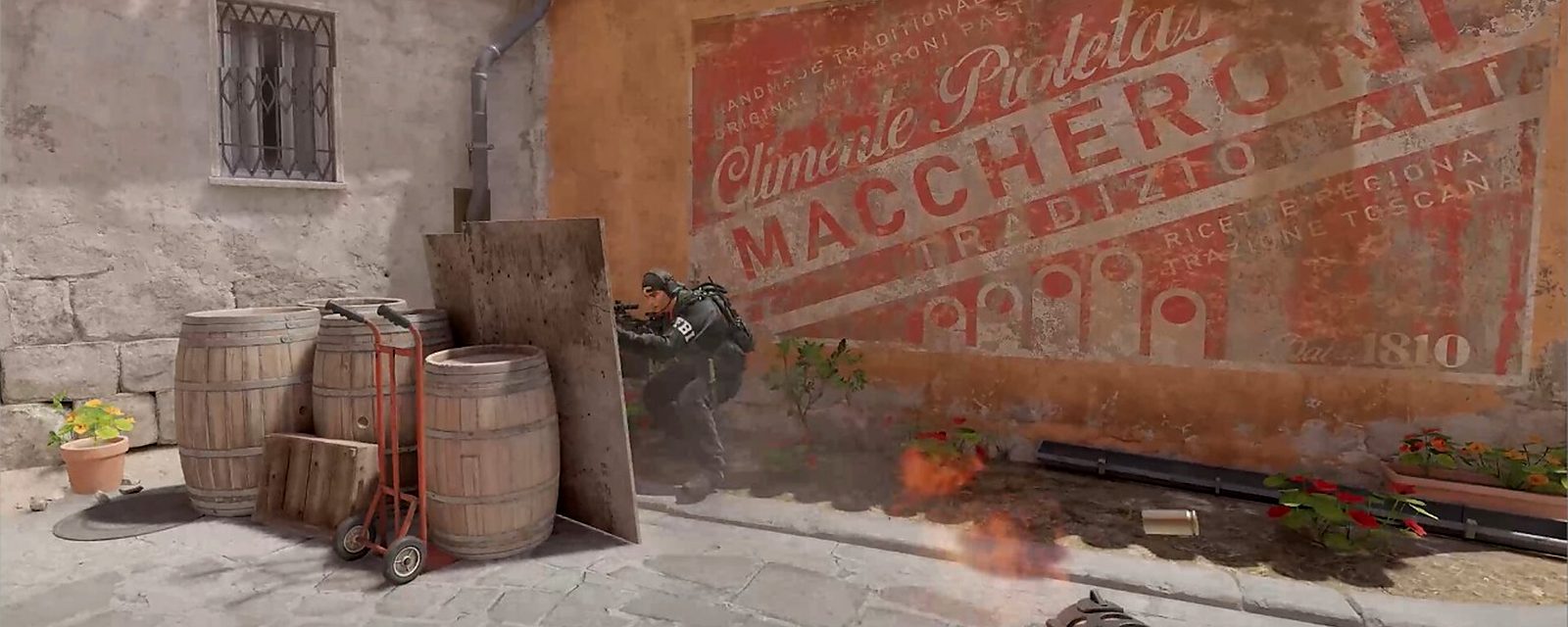 A CS2 player firing from behind barrels beside the painted Maccheroni wall on Inferno