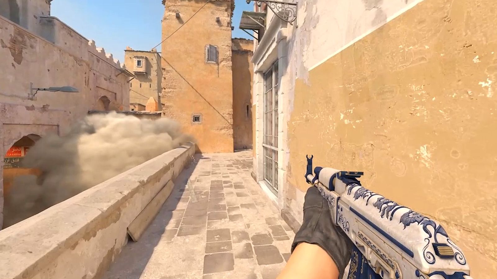 Counter-Strike 2 first-person view on Dust II with an AK-47 held on the right as a smoke rises behind a wall